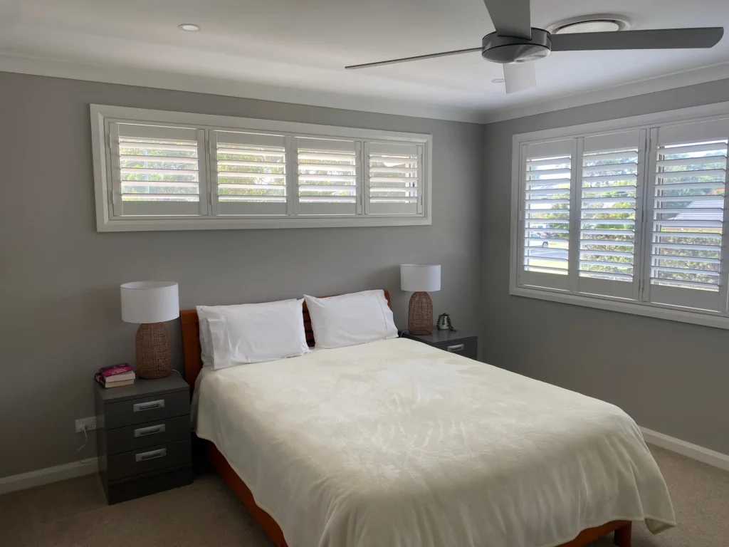 Modern bedroom with soft grey painted walls, white plantation shutters, ceiling fan, and neatly made bed with matching bedside tables.